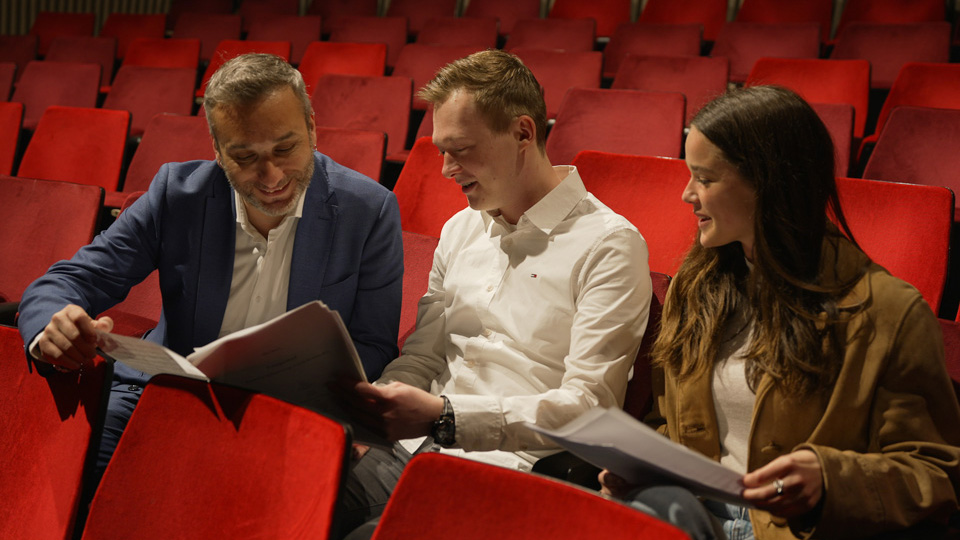 A man wearing a suit talking to a male and female students, looking at their music sheets, sitting in red theatre chairs.